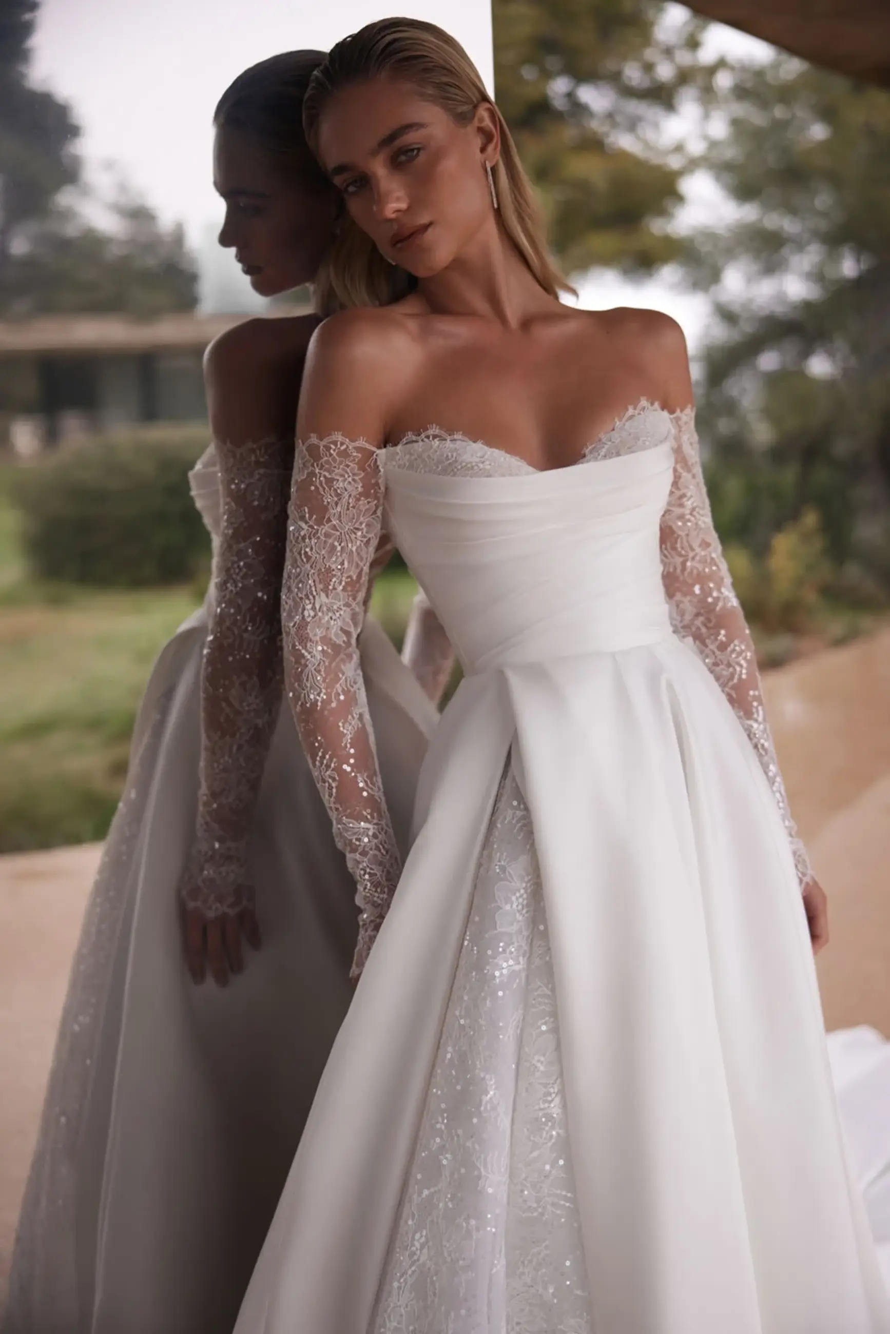 A woman in an elegant off-shoulder white wedding dress with lace sleeves stands by a reflective surface outdoors, conveying a serene and sophisticated mood.