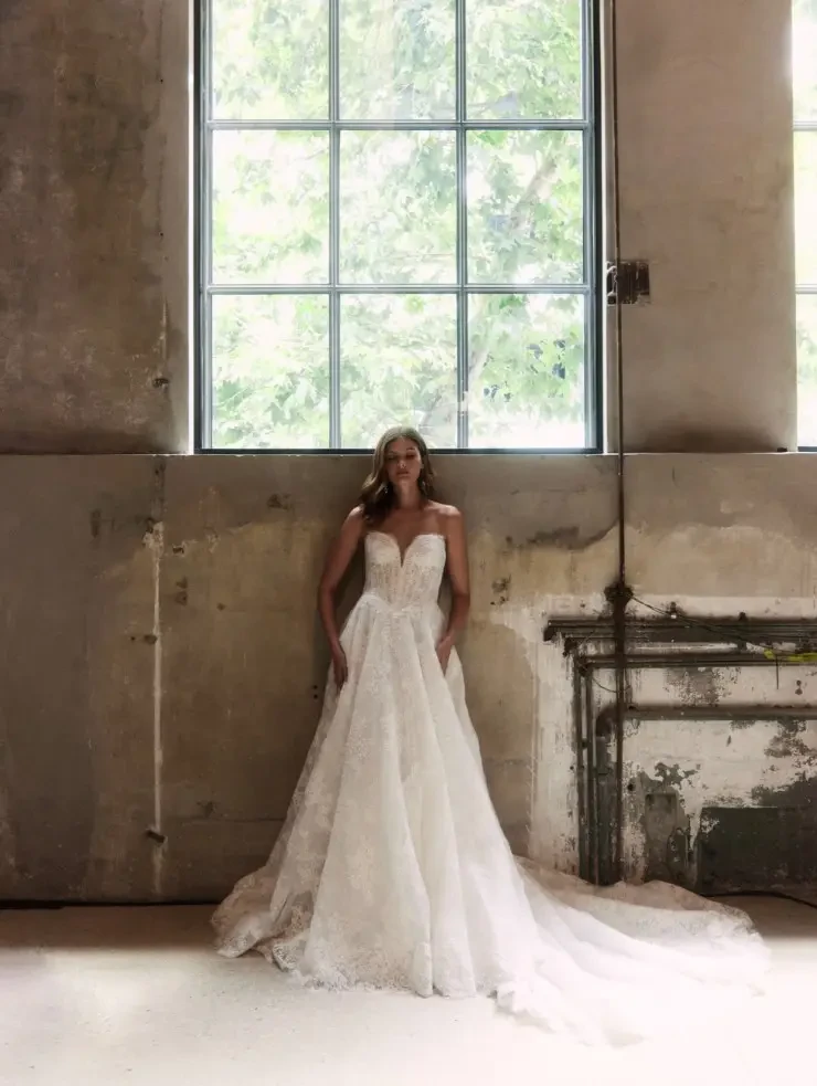 Model is wearing a chantilly lace A-line gown with a dramatic basque waist, elongated v-neckline, and optional mantilla veil.