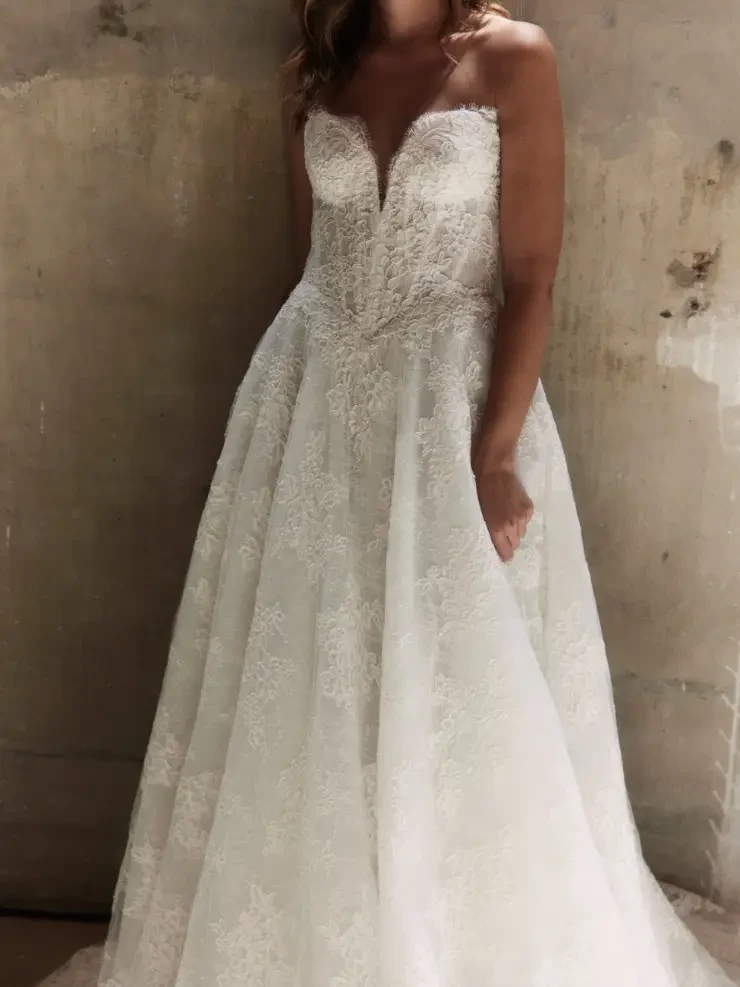 Model is wearing a chantilly lace A-line gown with a dramatic basque waist, elongated v-neckline, and optional mantilla veil.