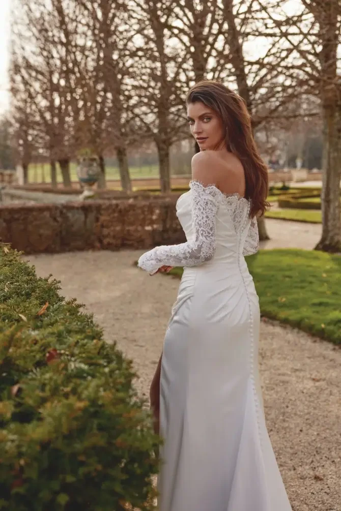 Model is wearing Jessa, a charmeuse fit-and-flare gown with an off-the-shoulder neckline and long lace sleeves.