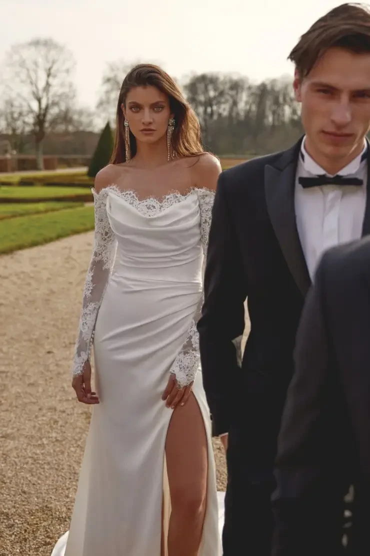 Model is wearing Jessa, a charmeuse fit-and-flare gown with an off-the-shoulder neckline and long lace sleeves.