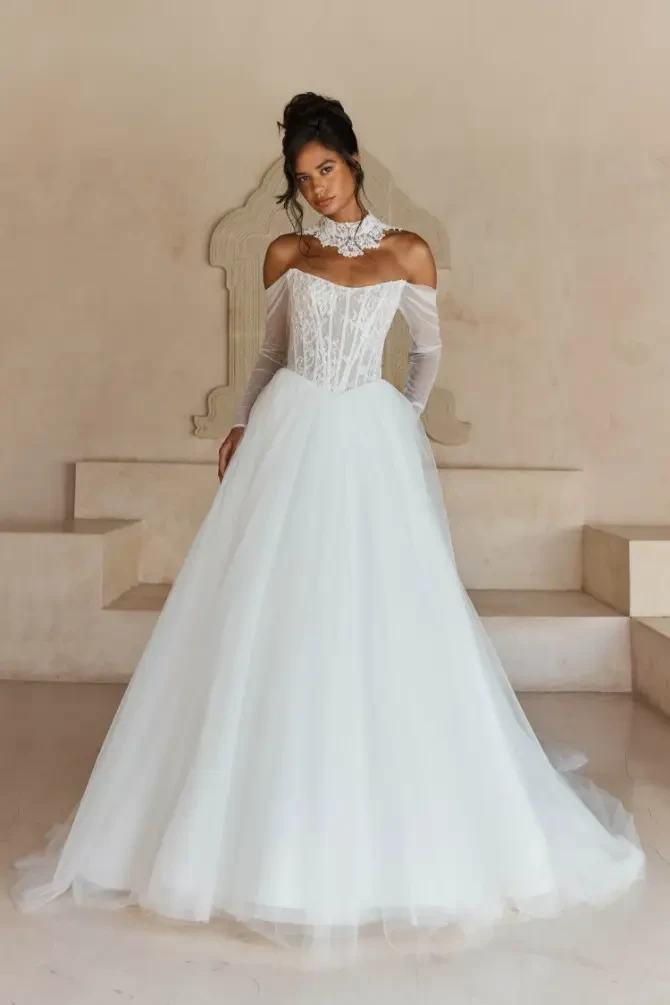 Model is wearing Larkin, a full tulle ball gown with a ruched long-sleeve bodice, beaded Chantilly lace, and optional beaded collar.