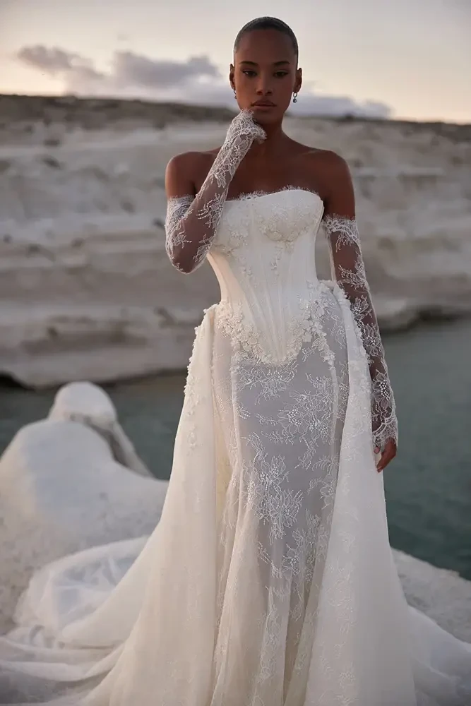 A woman in an elegant, off-the-shoulder white lace gown with long sleeves stands on a rocky shore at sunset, overlooking calm blue water, exuding tranquility.