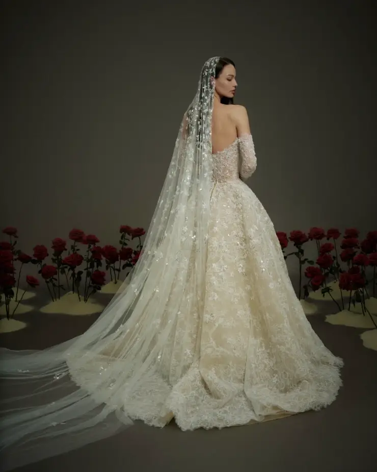 Model is wearing NF108, a ball gown with a sweetheart neckline, off-the-shoulder sleeves, a sheer sequined corset bodice, and a full skirt with matching cathedral-length veil.