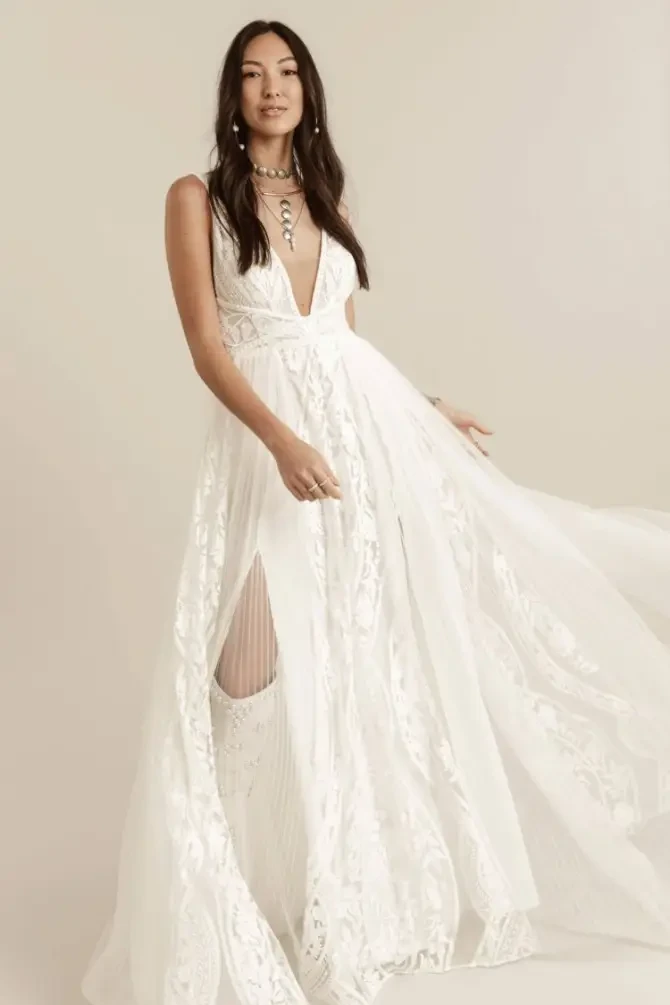 Model is wearing a sleeveless a-line wedding dress that has pleated tulle panels and floral embroidery, with a plunging neckline and double slit 