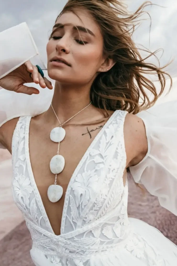 Model is wearing a sleeveless a-line wedding dress that has pleated tulle panels and floral embroidery, with a plunging neckline and double slit 