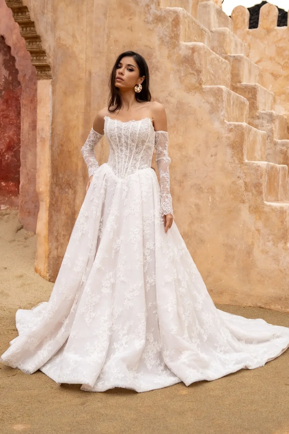 Woman in elegant off-shoulder lace wedding dress poses outdoors against textured stone wall with arches and steps. The tone is serene and sophisticated.