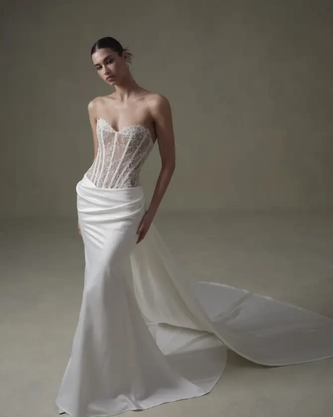 Model is wearing a sleek strapless fit and flare gown with exposed boning, a lace-covered bodice, and a smooth, dramatic train