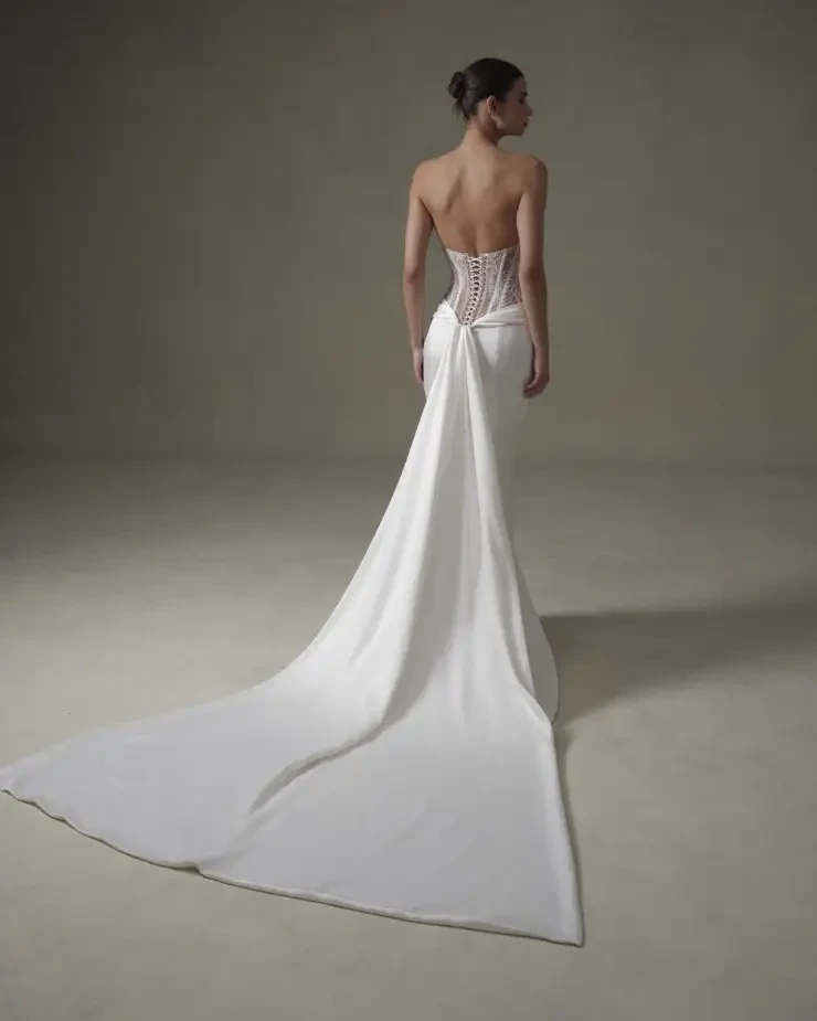 Model is wearing a sleek strapless fit and flare gown with exposed boning, a lace-covered bodice, and a smooth, dramatic train
