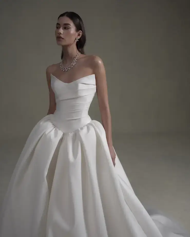 A woman in an elegant white strapless gown with a full, flowing skirt poses confidently. She wears a statement necklace, exuding sophistication and grace.