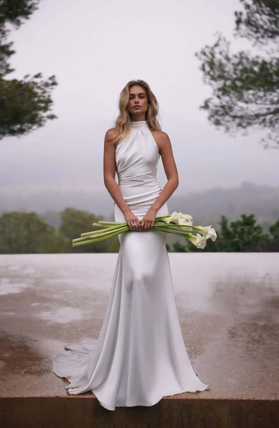 A woman in an elegant, white, halter-neck gown holds calla lilies, standing on a wet stone surface surrounded by blurred greenery, creating a serene atmosphere.