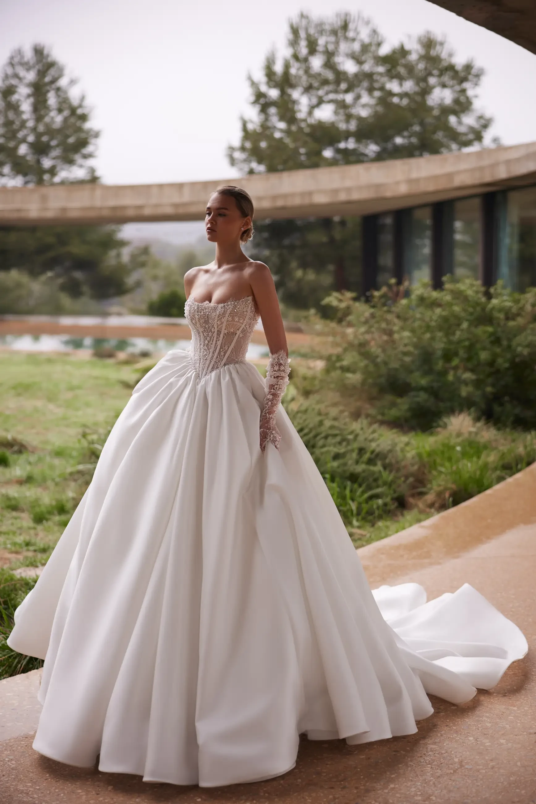 Bride in an elegant, strapless white gown with a voluminous skirt, standing outdoors. The setting feels serene and natural, enhancing elegance.