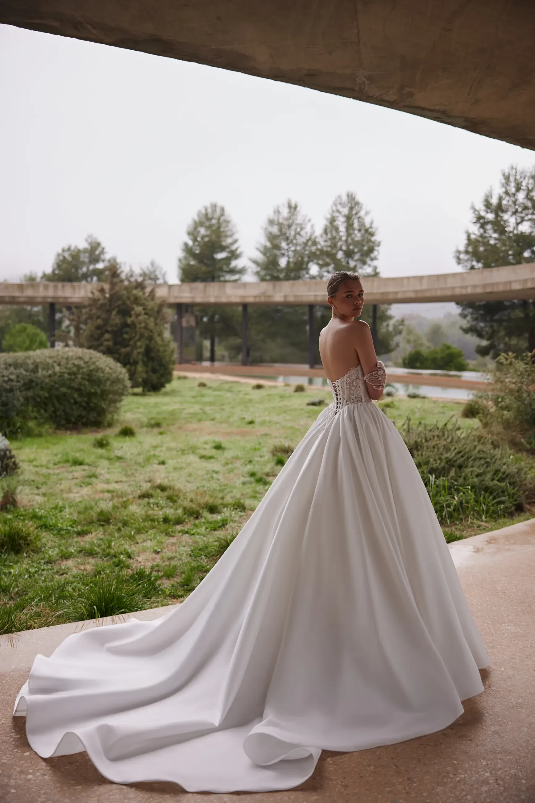 Bride in strapless white gown with long train stands outdoors on a misty day. Lush greenery and modern architecture create a serene, elegant scene.