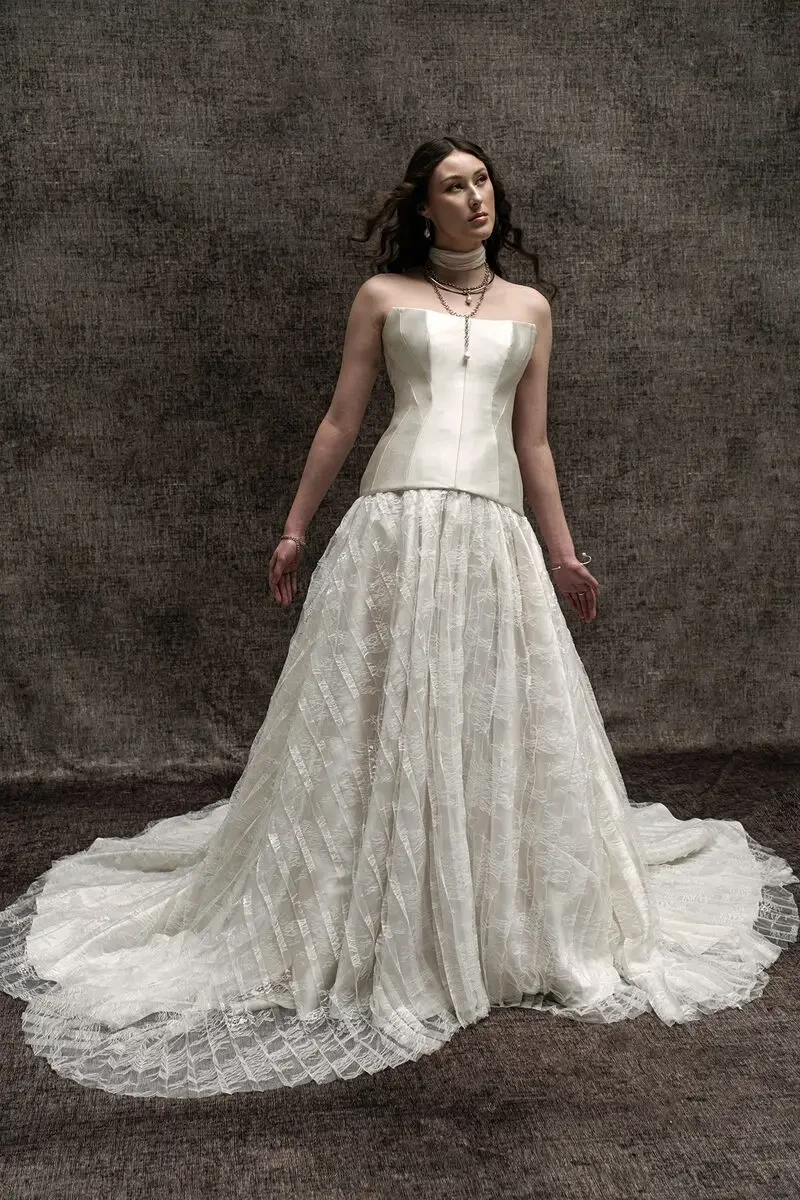 A woman in a vintage, elegant wedding dress standing against a textured background. The dress features a fitted bodice and a flowing, layered skirt.