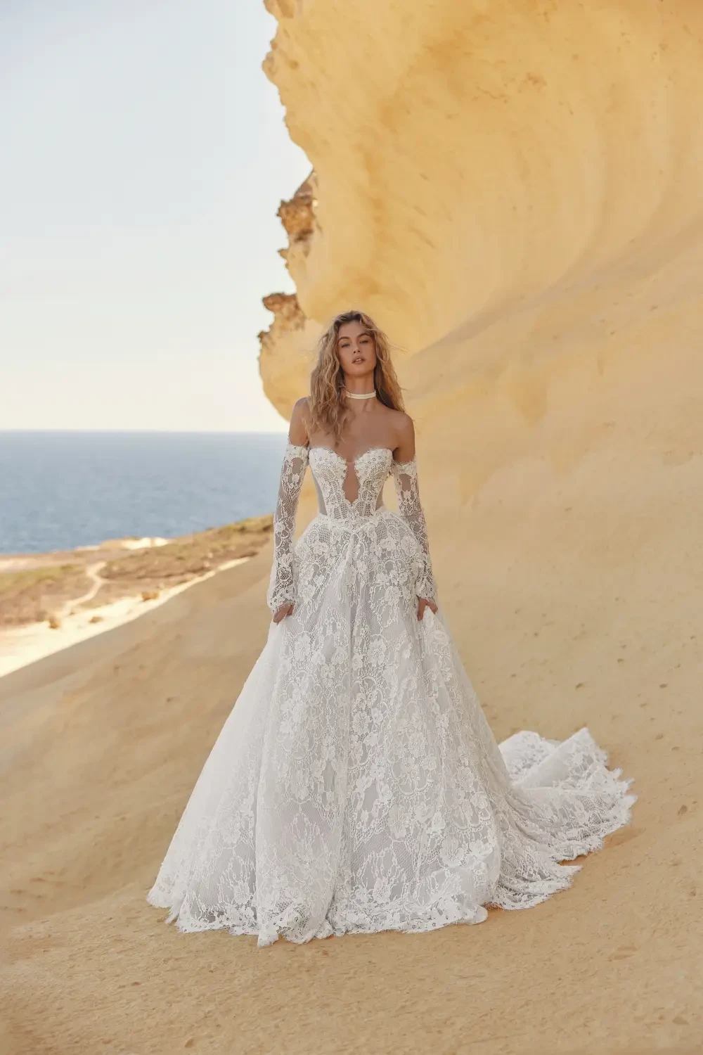 A woman in an elegant, intricately designed white lace wedding gown stands on a sandy cliff overlooking the ocean.