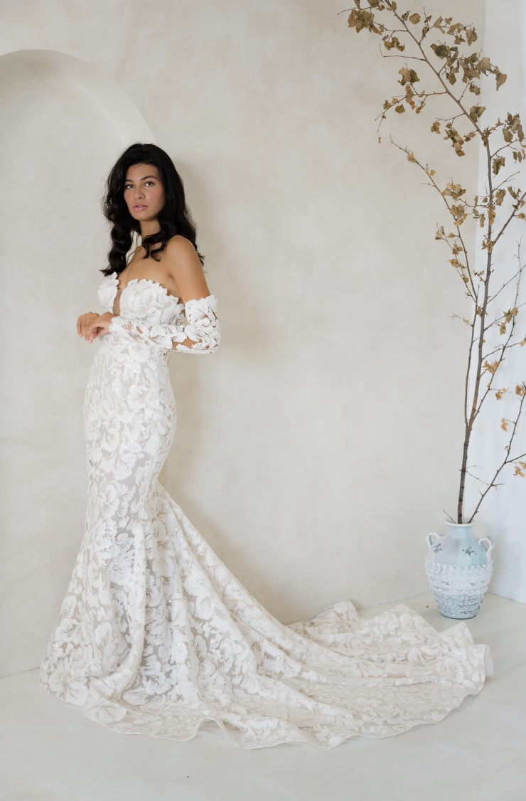 Woman in an elegant, off-the-shoulder lace wedding gown stands in a minimalist room. A tall, delicate branch in a vase adds a touch of nature.