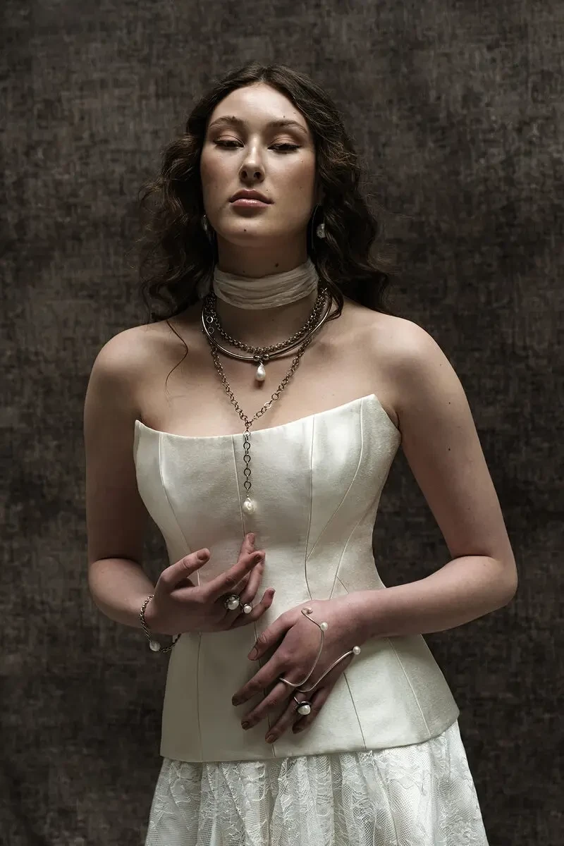 A woman in a white corset dress with layered necklaces poses confidently.