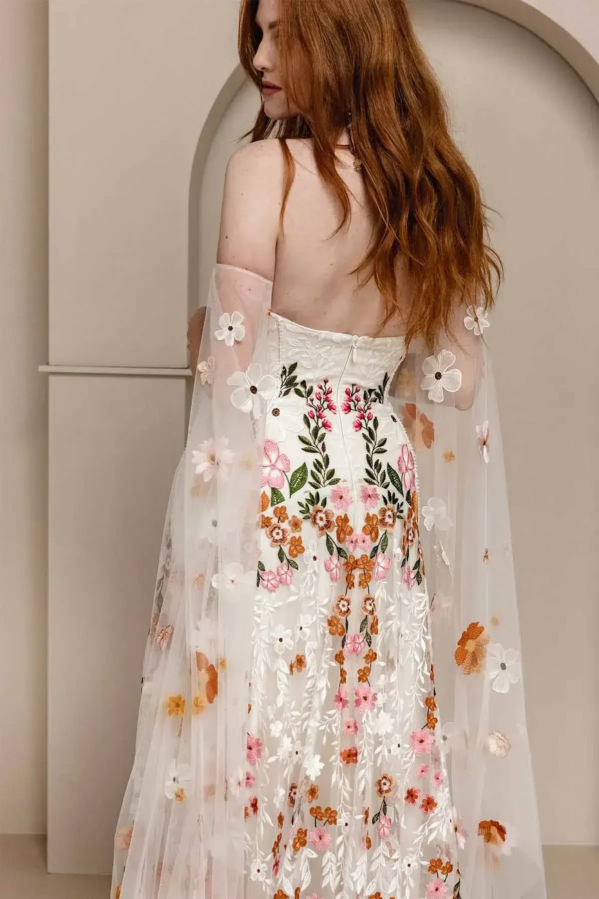 Model is wearing a sand-dyed tulle gown with vibrant floral embroidery, a structured boned bodice, and a dramatic floral train.