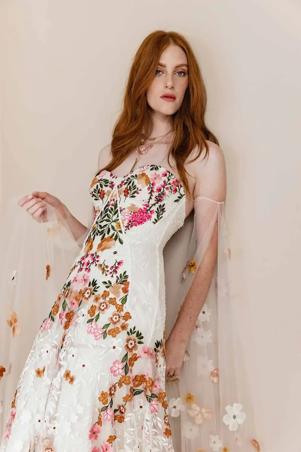 Model is wearing a sand-dyed tulle gown with vibrant floral embroidery, a structured boned bodice, and a dramatic floral train.