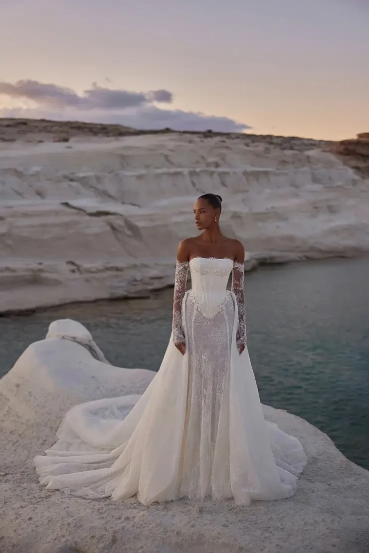 A woman in an elegant, off-the-shoulder white lace gown with long sleeves stands on a rocky shore at sunset, overlooking calm blue water, exuding tranquility.