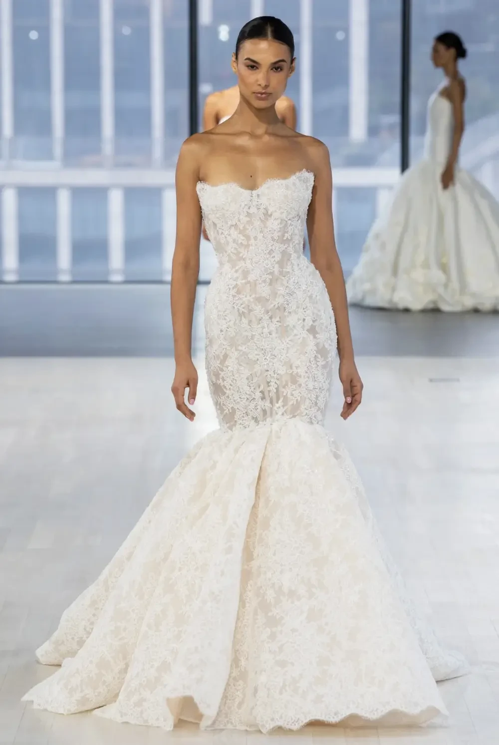 A model wearing a strapless, white mermaid wedding gown with intricate lace detailing, walking down a runway.