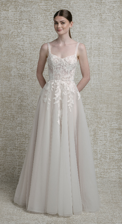 A woman stands wearing a delicate, floor-length wedding dress with intricate floral embroidery and thin straps, against a textured neutral background.