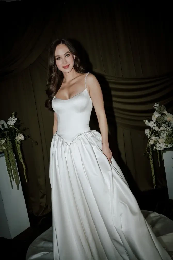 A woman in a white gown poses elegantly against a dark backdrop with floral arrangements beside her.