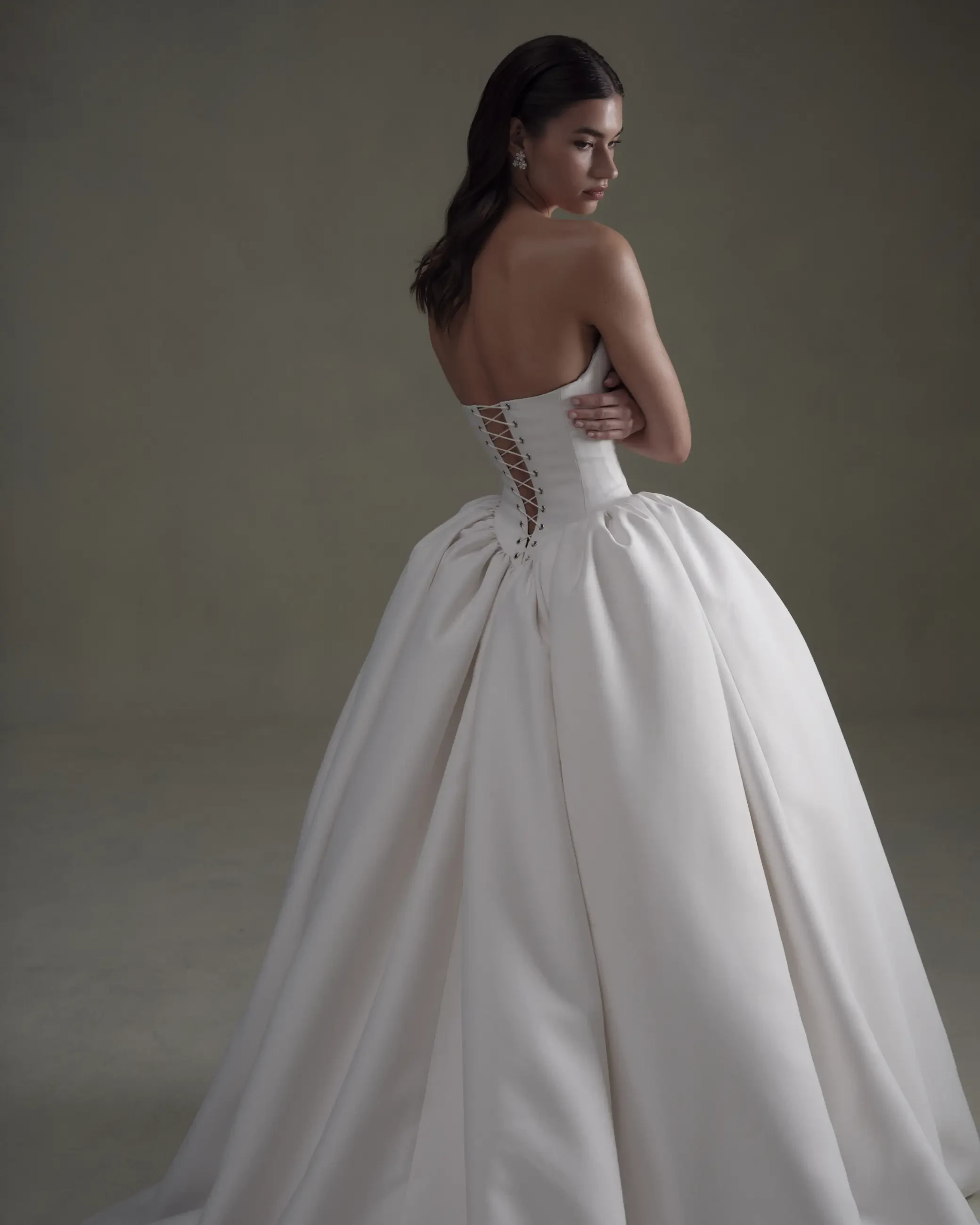 A woman in an elegant white strapless ball gown, featuring a lace-up back, stands against a neutral background. She exudes grace and sophistication.