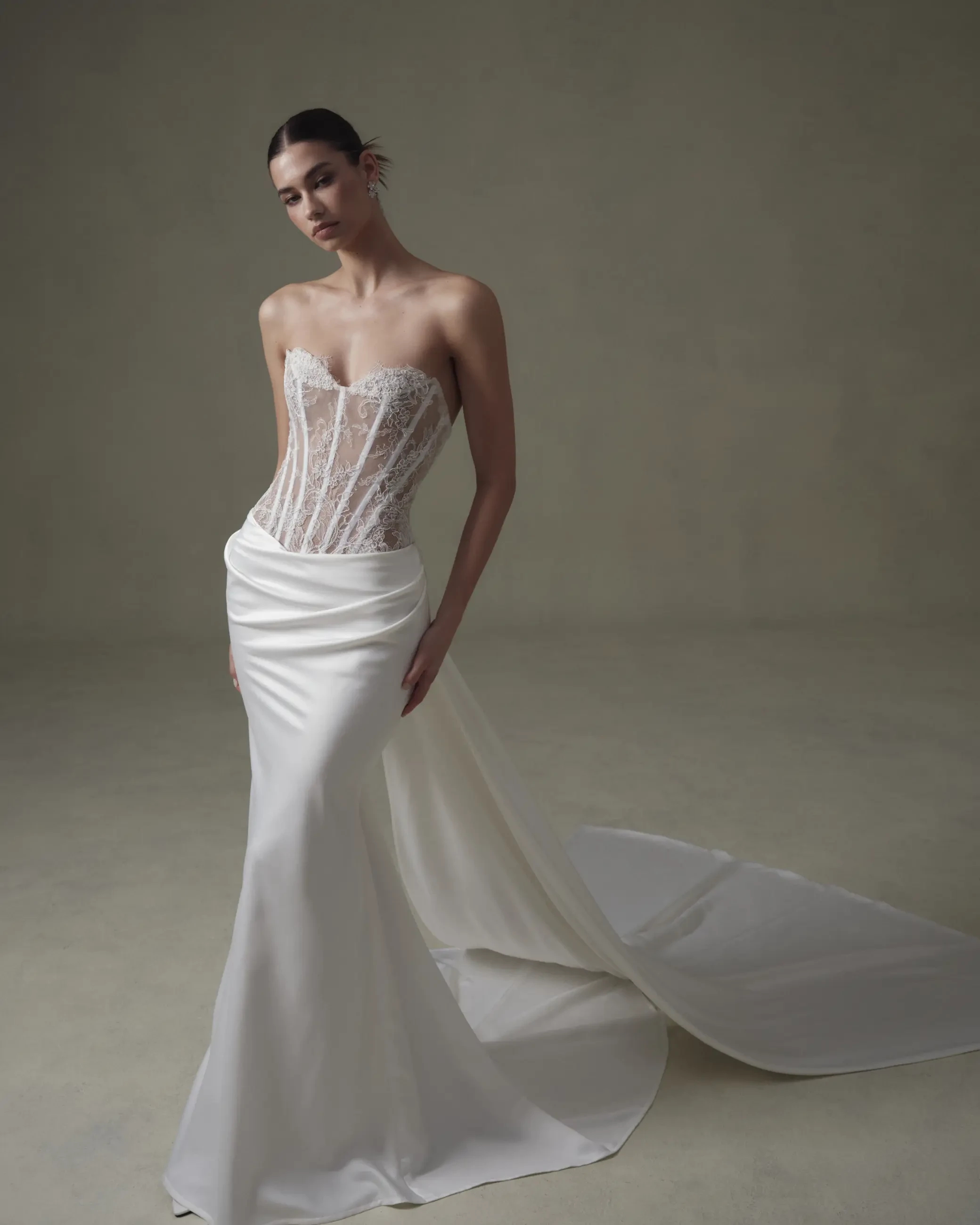 A woman in an elegant, strapless white gown with a lace corset bodice and flowing satin skirt stands gracefully against a soft, muted background.